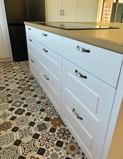 Nowoczesna kuchnia z jasną wyspą, drewnianym blatem i białymi szufladami z ceramicznymi uchwytami retro.